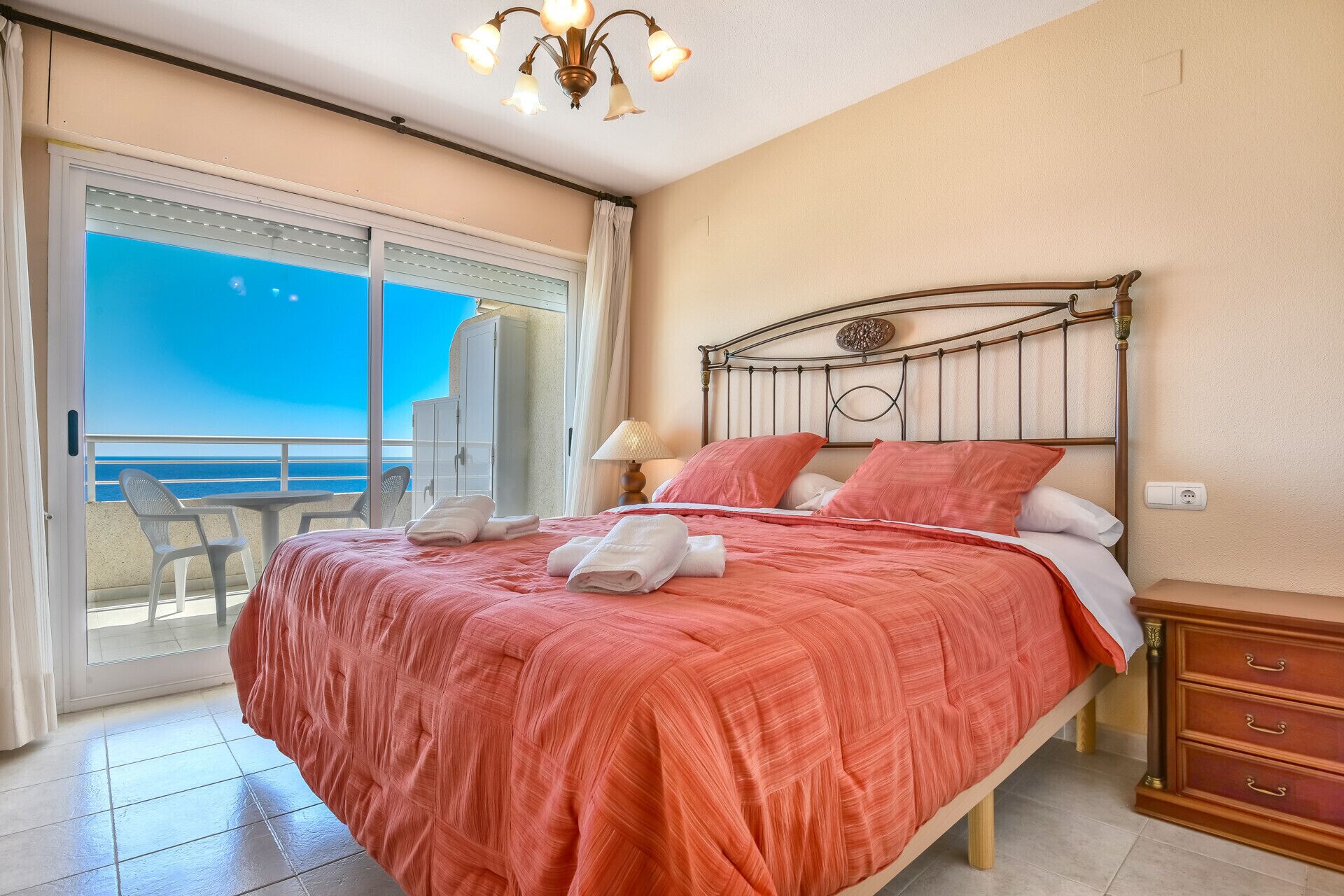 A bedroom with a bed, a nightstand, a lamp, a wardrobe, and a view of the sea through a sliding door.