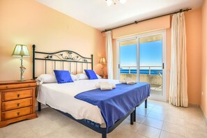 Un dormitorio con cama, dos mesitas de noche, un aparador de madera y un balcón con vistas al mar.