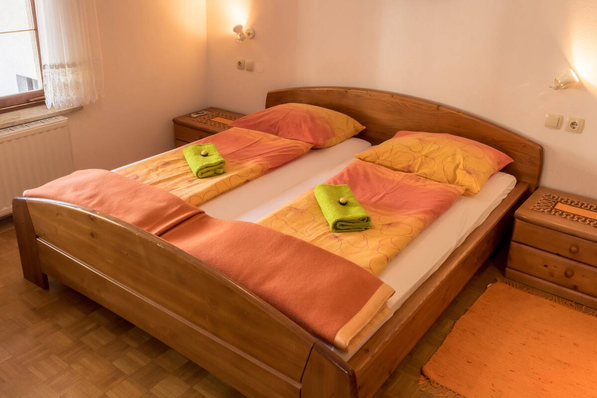 Cama doble con cabecera y piecera de madera, dos almohadas y una toalla verde a cada lado.
