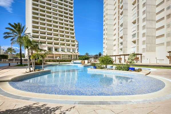 A swimming pool surrounded by a landscaped area with palm trees, adjacent to high-rise buildings.