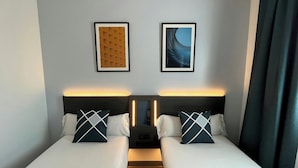 A hotel room with two beds, each with a pillow and a headboard. There are two framed pictures on the wall, a nightstand with a lamp, and a wooden floor.