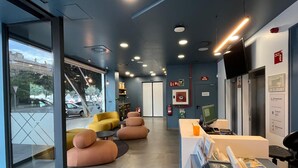 A modern reception area with a glass door leading outside, a seating area with cushioned chairs, a reception desk with a computer, and a wall-mounted TV.