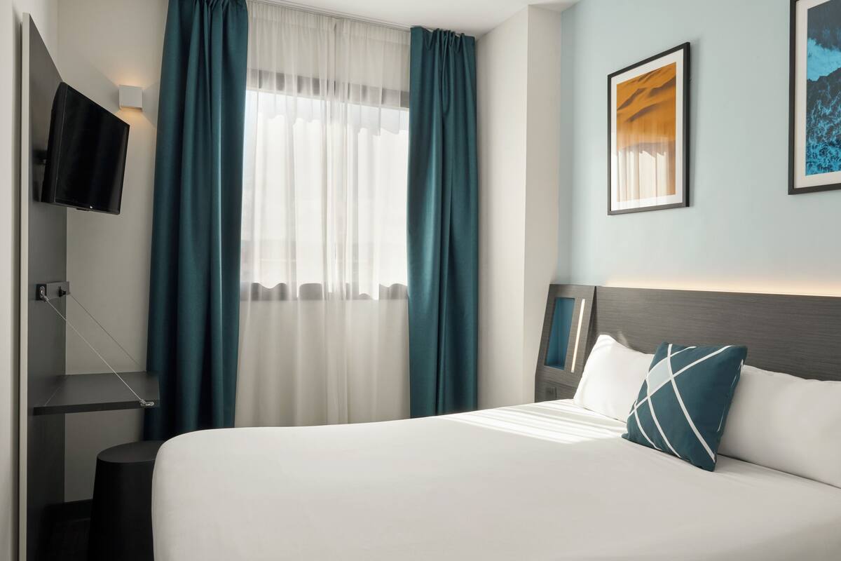 Habitación de hotel con cama, televisor montado en la pared, ventana con cortinas e imagen en un marco colgado en la pared.