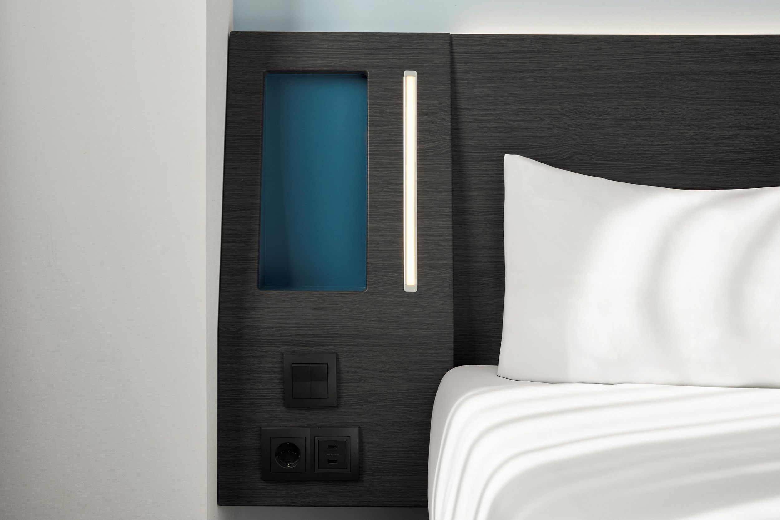 A modern hotel room with a dark wood headboard, a bedside table with a blue panel, a white pillow, and a power outlet.