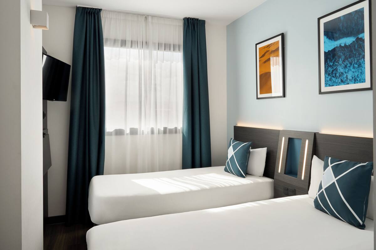 Habitación de hotel con dos camas, un televisor, una ventana con cortinas e imágenes enmarcadas en la pared.