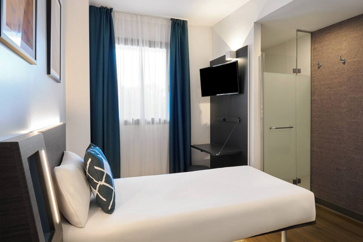 Una habitación de hotel moderna con cama, televisión montada en la pared, zona de ducha y ventana con cortinas.