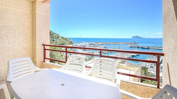 Un balcon avec une table et des chaises, donnant sur une marina où sont amarrés des bateaux, avec un ciel bleu dégagé.