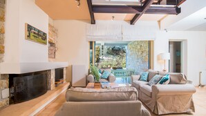 A cozy living room with a fireplace, a sofa set, and a view of the garden. - Villa Lithia Ano Korakiana (Ano Korakiana)