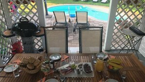 Mesa de comedor dispuesta con platos, cubiertos y diversos alimentos, con vista a una piscina.