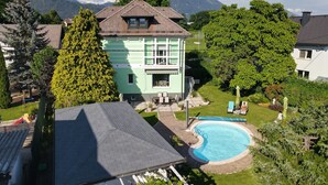 Una casa de dos pisos con piscina, rodeada de abundante vegetación y montañas al fondo.