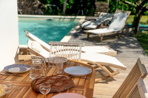 A wooden table set with plates, glasses, a pitcher, and a loaf of bread, overlooking a pool and lounge chairs.