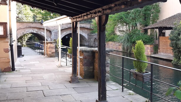 A stone pathway leading to a bridge over a canal, with a wooden pergola and a small building with a tiled roof in the background. - "LANDBeautiful beamed apartment in the historic center of Colmar's "Little Venice". (Colmar)
