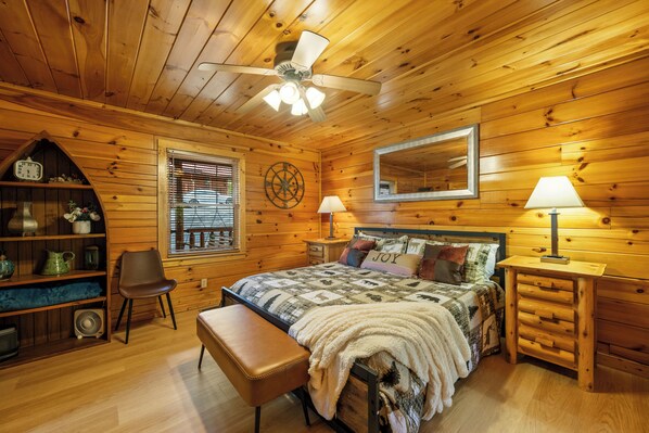 Une chambre confortable dans un chalet en bois, avec un lit, un banc, un ventilateur, une lampe, un miroir et une fenêtre avec des stores.