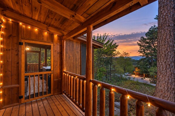 A wooden cabin with a balcony, string lights, and a view of the surrounding forest.