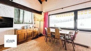 Una habitación acogedora con un armario de madera, una mesa con sillas, un ventanal con vistas a árboles y una lámpara.