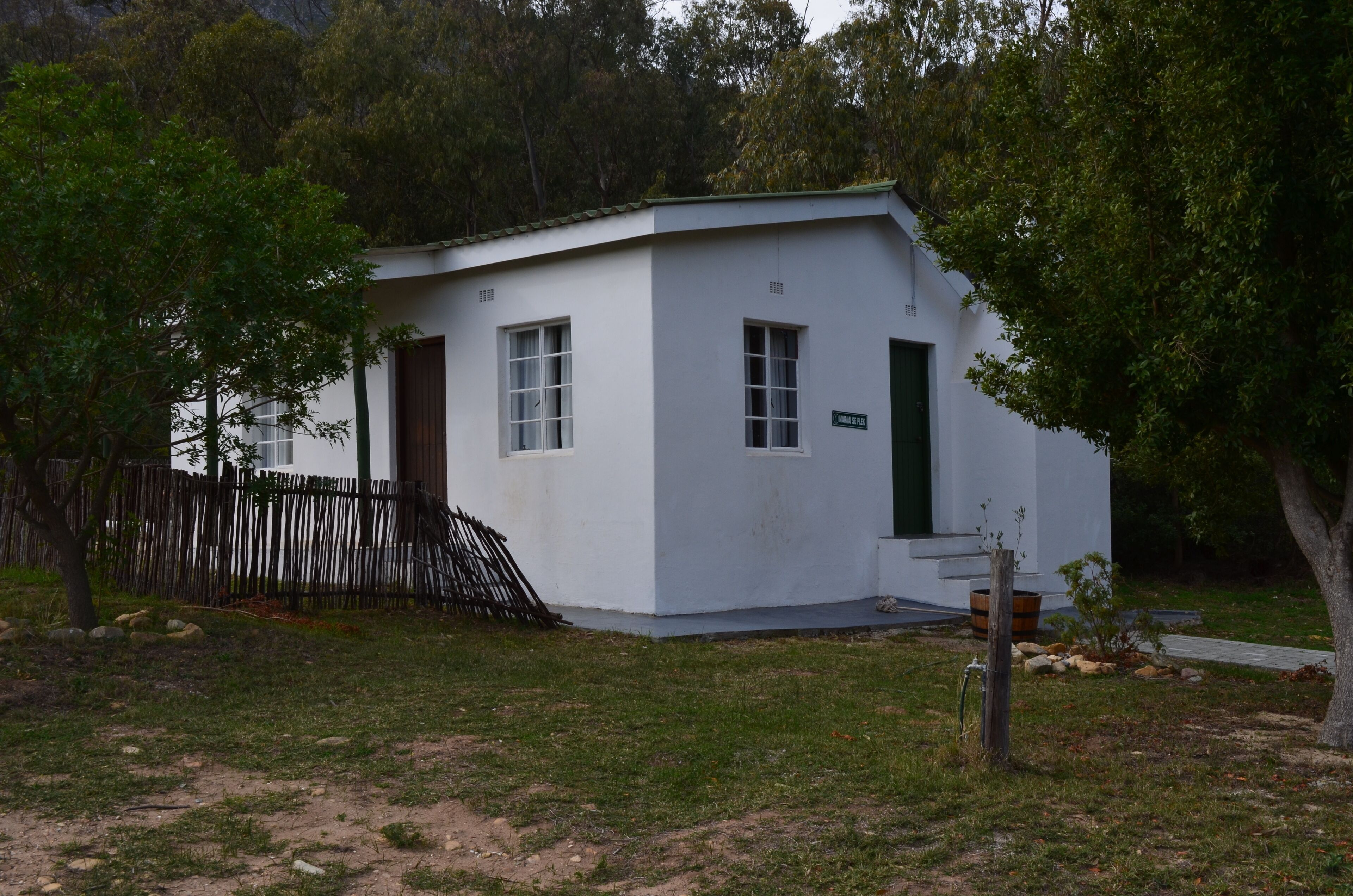 A small, white building with a green door and windows, surrounded by trees and a wooden fence.