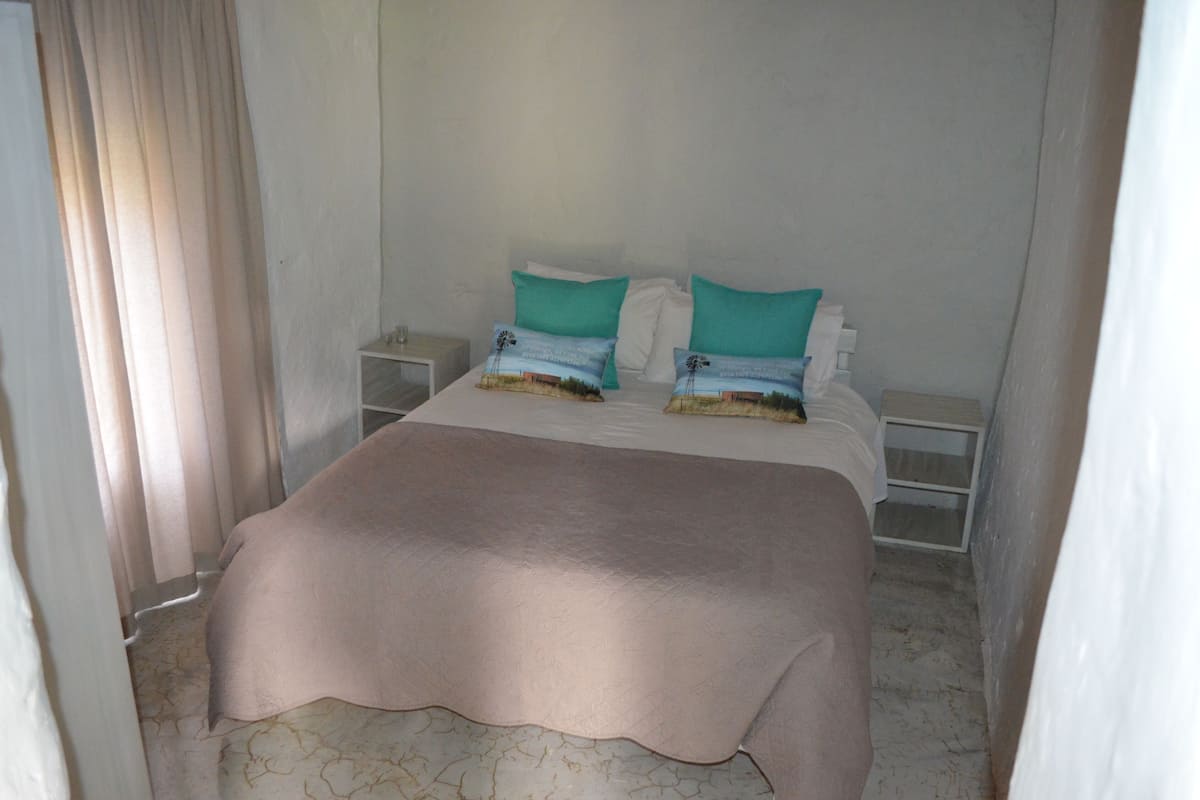 A neatly made bed with a beige bedspread and two teal pillows, a bedside table with a lamp, and a curtain partially drawn to one side.