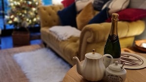 A cozy living room with a Christmas tree, a sofa with cushions, a table with a teapot and a bottle, and a large window with a view. - Romantic streamside Carriage next to Coast Path ♥️ Short walk to beach & pub! 🥂 (Ruan minor)
