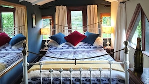 A bed with a white metal frame and a patterned bedspread, a wooden nightstand with a lamp, and a window with curtains. - Romantic streamside Carriage next to Coast Path ♥️ Short walk to beach & pub! 🥂 (Ruan minor)
