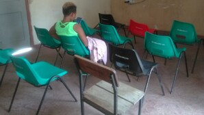 A room with a television mounted on the wall, a person sitting on a chair, and several other chairs arranged around a table. - Murbs Hostel (Kisumu)