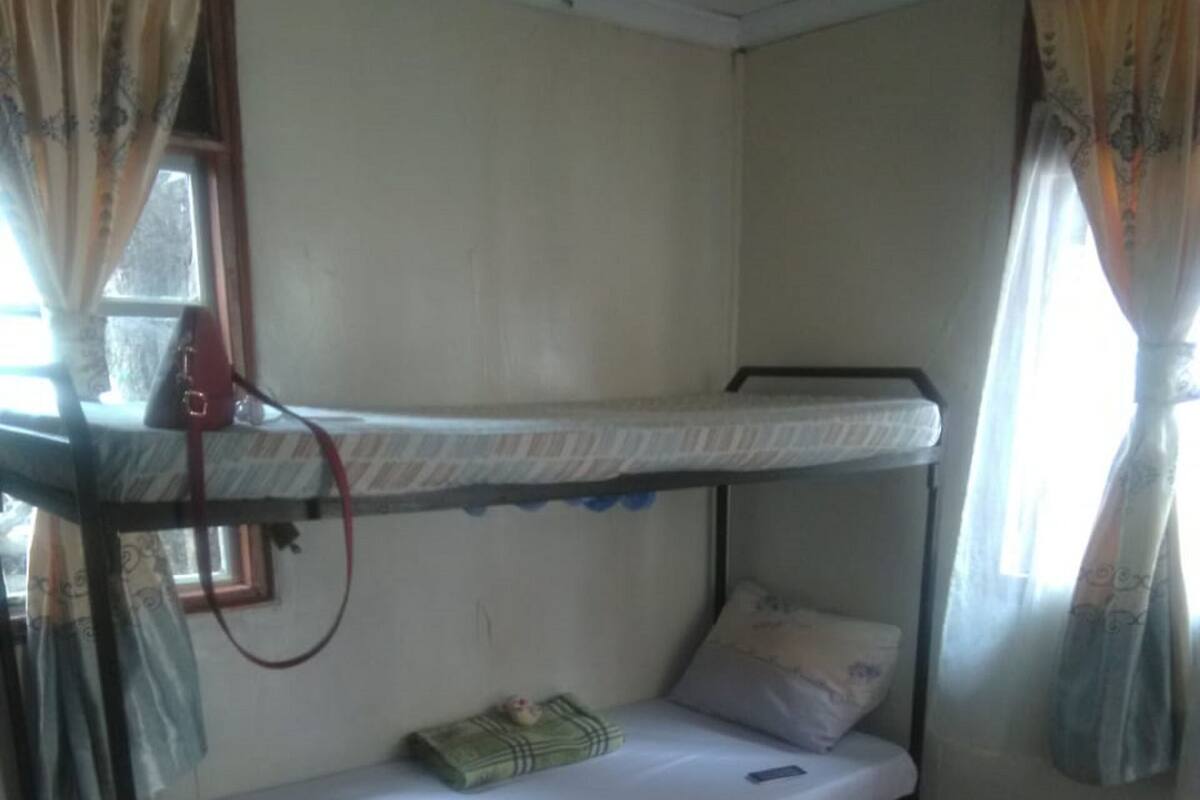 A small, single-bunk room with a patterned floor, a red rug, and curtains on the windows.