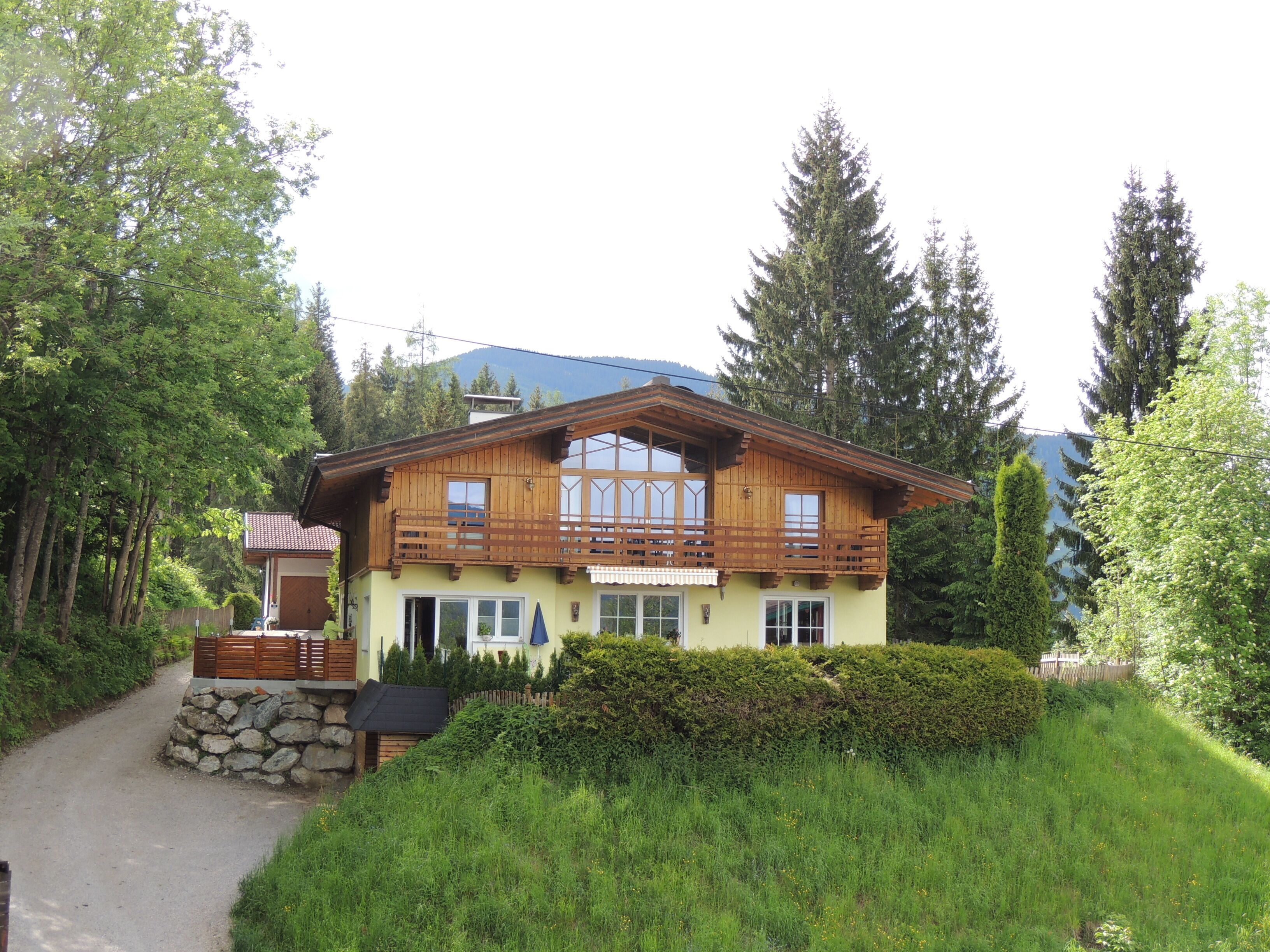 A two-story wooden house with a balcony, surrounded by greenery and trees.