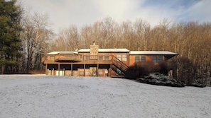 Een houten chalet met twee verdiepingen en een balkon, omgeven door een besneeerd landschap en dichte bomen.
