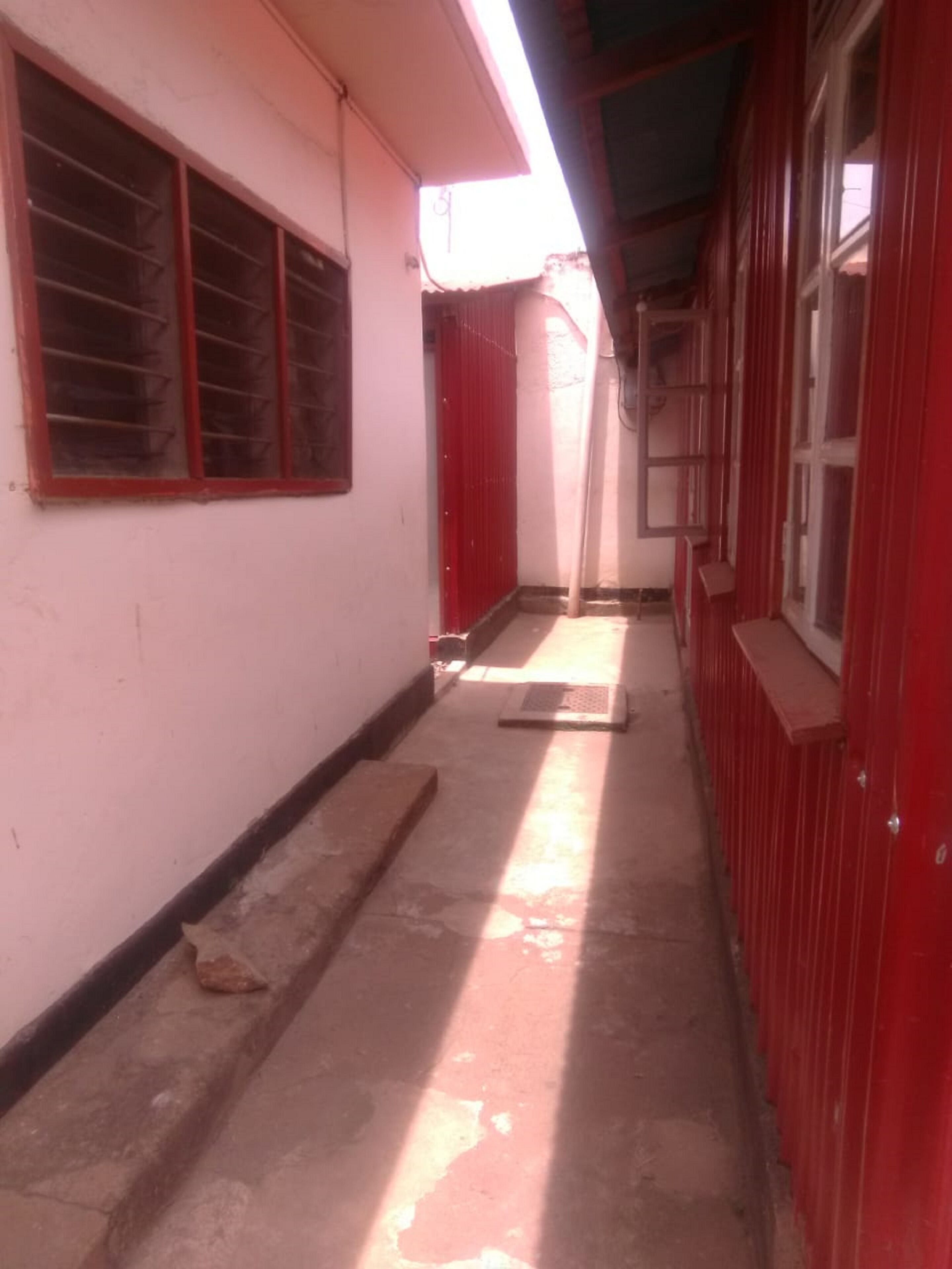 Un corredor estrecho con una puerta roja y ventanas, que da a una habitación con suelo de hormigón y una puerta de metal.