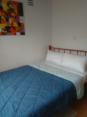 A single bed with a blue quilt and white pillows, a red headboard, and a colorful abstract painting on the wall.