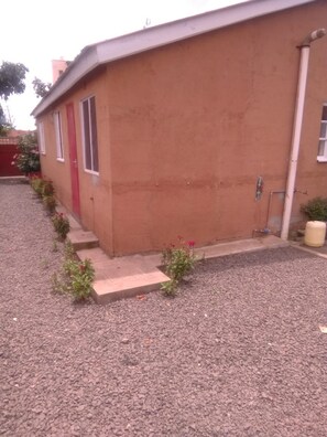 A single-story building with a red door and windows, a gravel driveway, and a small garden area with red flowers.