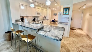 A modern kitchen with white cabinets, a granite countertop, and pendant lighting. There are two stools and a trash can.