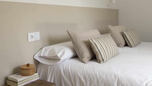 Una cama bien hecha con ropa blanca y almohadas a rayas, una mesita de noche con libros y un recipiente de madera, y un enchufe de pared.