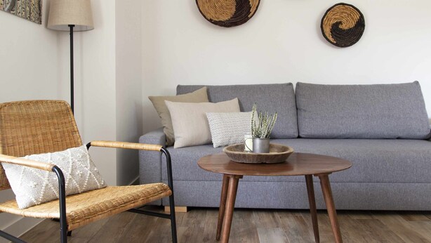 Un salón con un sofá gris, una silla de mimbre, una mesa redonda de madera y una lámpara de pie.