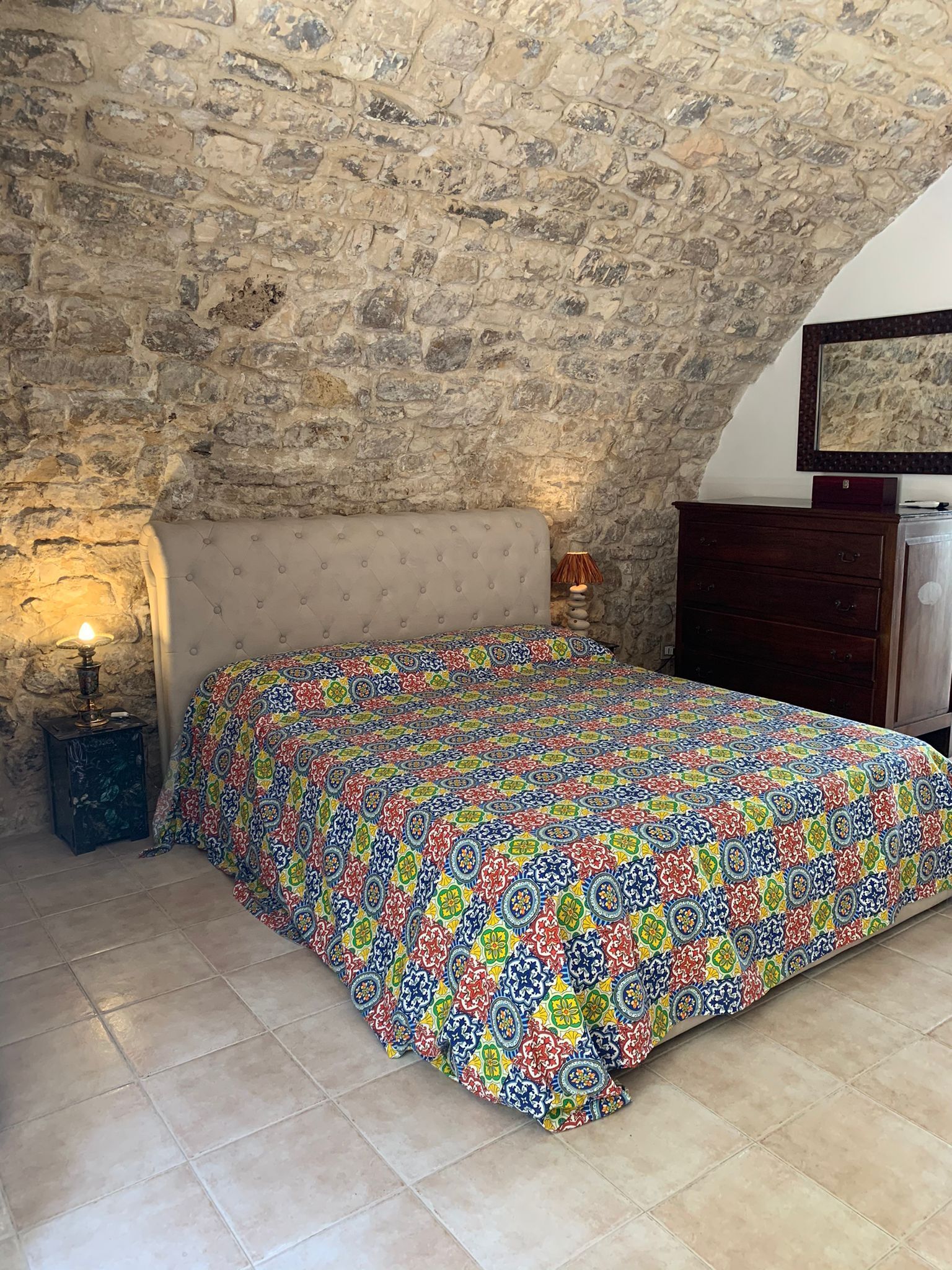 Un dormitorio con pared de piedra, una cama con colcha de motivos coloridos, una lámpara de noche y un aparador.