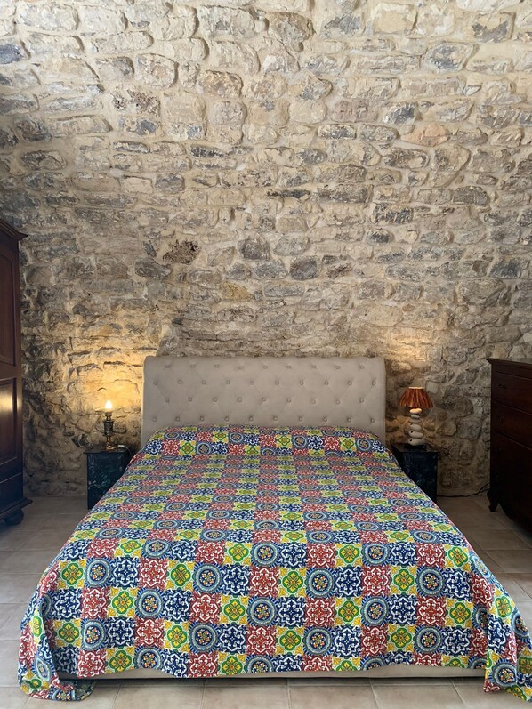 Un dormitorio con pared de piedra, cabecera capitoneada, colcha estampada y dos mesitas de noche con lámparas.