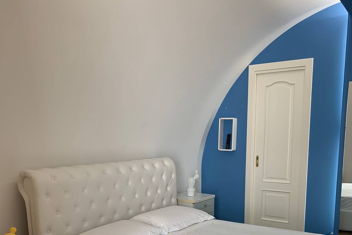 Un dormitorio con una cabecera tapizada en blanco, una mesita de noche de madera, una colcha blanca y una pared de acento azul.