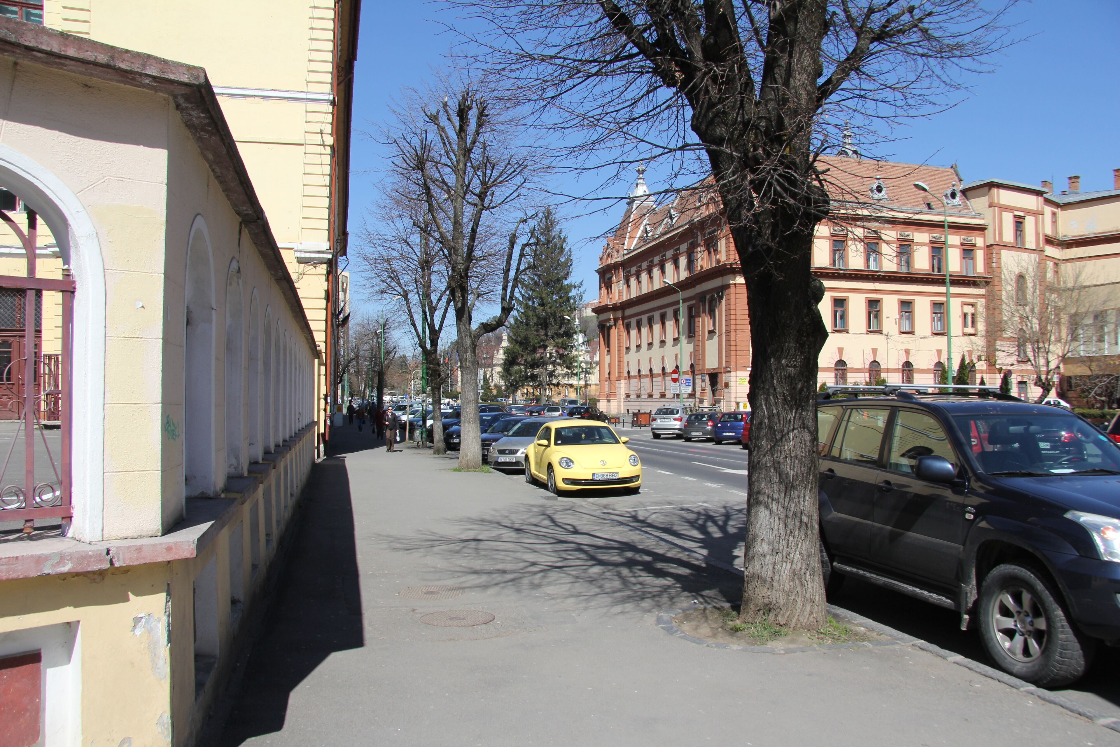 A street with parked cars, a yellow car in the foreground, and buildings with arched windows on the left.
