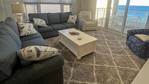 Ein Wohnzimmer mit einer dunkelgrauen Couch, einem weißen Couchtisch und einem gemusterten Teppich. Es gibt einen Deckenventilator, ein Fenster mit Jalousien und einen Blick auf den Ozean durch große Glasüren.