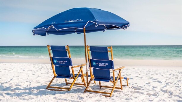 Zwei blaue Liegestühle mit 'La Dolce Vita'-Branding und ein passender blauer Sonnenschirm an einem Sandstrand.