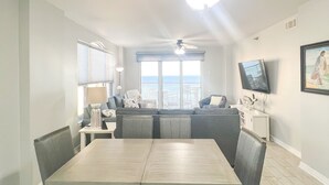 Ein Essbereich mit Tisch, Stühlen und Deckenventilator. Im Hintergrund befindet sich eine Couch und ein Fernseher.