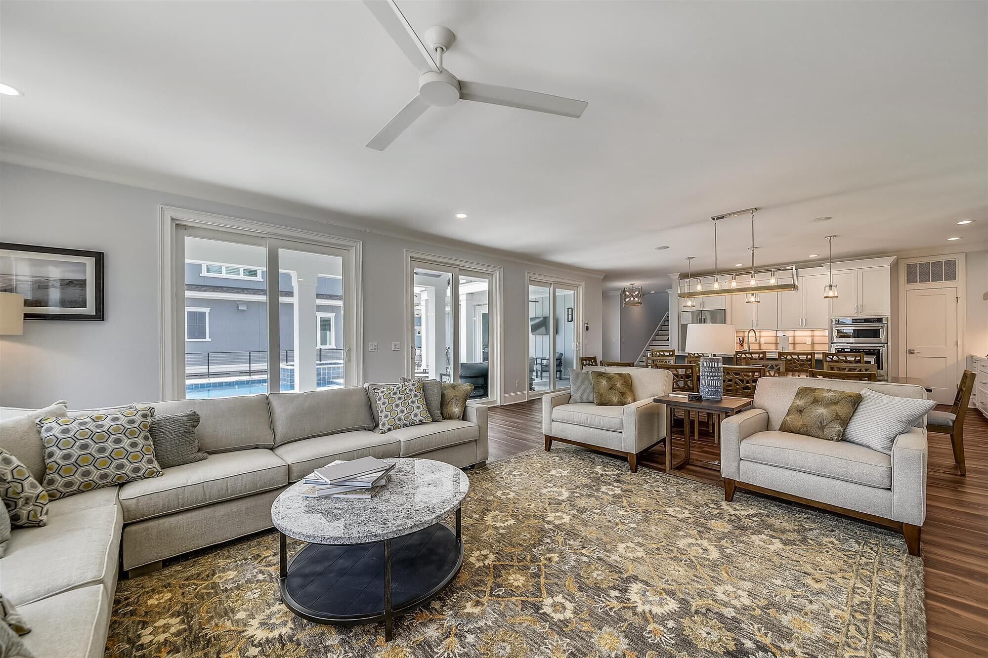 A spacious living room with a grey sofa, a round coffee table, and a patterned rug. There is a dining area with wooden chairs and a chandelier.