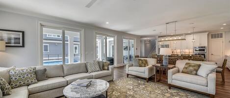 A spacious living room with a grey sofa, a round coffee table, and a patterned rug. There is a dining area with wooden chairs and a chandelier.