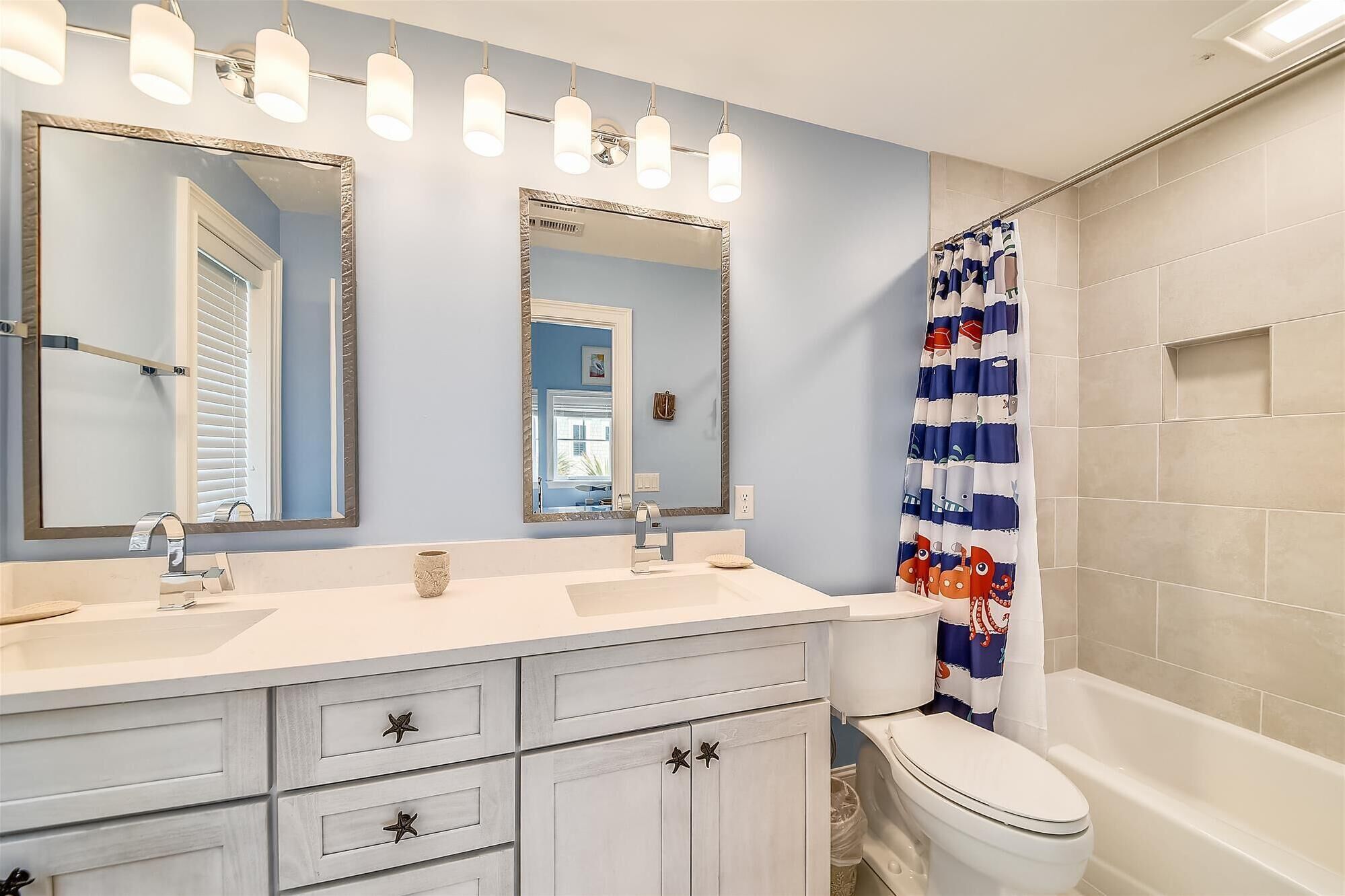 A bathroom with two sinks, a toilet, and a bathtub. There are three mirrors above the sinks, and a shower curtain with a fish pattern.