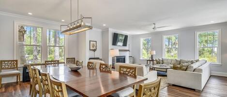 A spacious living room with a wooden dining table, comfortable seating, a fireplace, and large windows offering a view of the outdoors.