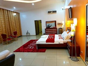 A hotel room with a bed, a desk, a chair, a red rug, a mirror, and a lamp. - Have'Den Resort-Hôtel (Abidjan)