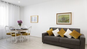 Una sala de estar con un sofá gris, sillas blancas, una mesa de comedor con un florero y dos cuadros en la pared.