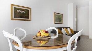 Un comedor con una mesa de vidrio redonda, sillas blancas y un cuenco de frutas. Hay dos obras de arte enmarcadas en la pared.