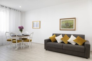 A living room with a grey sofa, white chairs, a dining table with a vase of flowers, and two framed pictures on the wall.