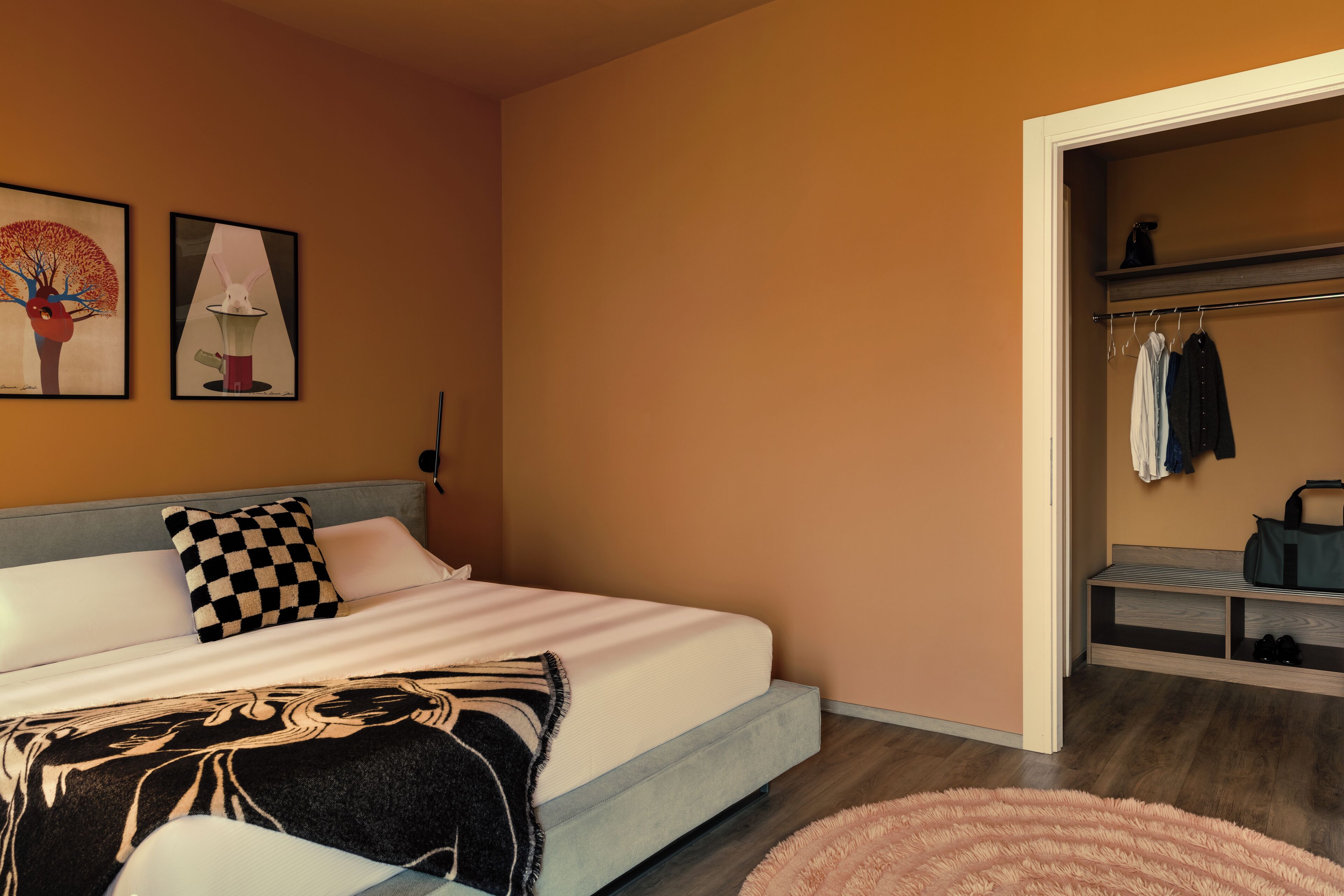 Une chambre à coucher avec un lit, un coussin à carreaux, une couverture, un tapis rose et une armoire contenant des vêtements et des chaussures.