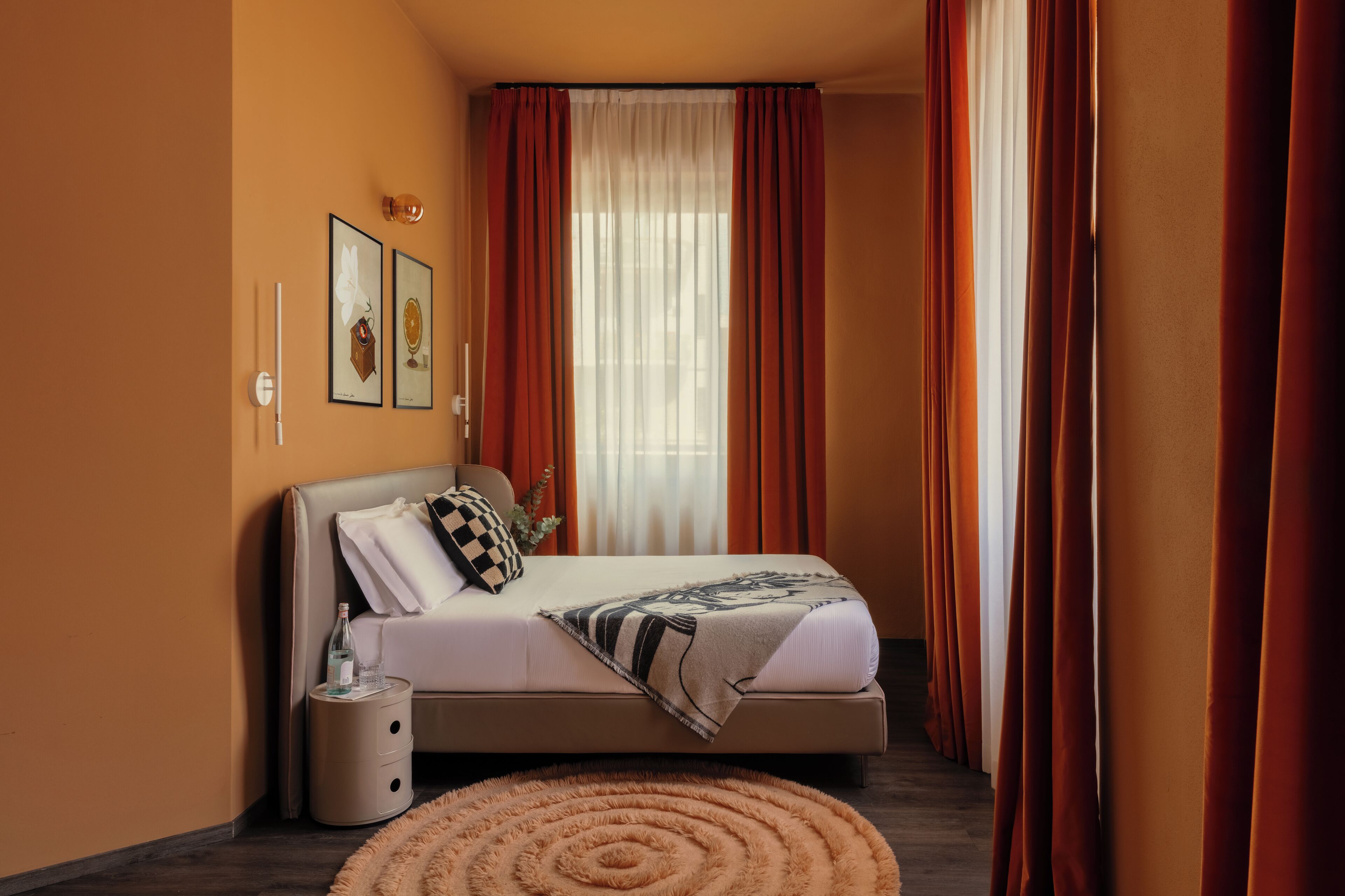 A bedroom with a bed, a bedside table, a round rug, and a window with curtains.